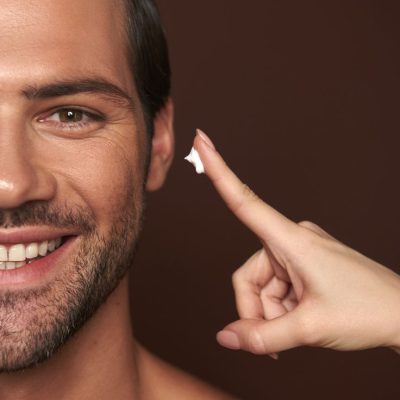 Beauty procedure and skin care. Close up half face portrait happy smiling handsome man looking at camera while female going to apply face cream on him. Isolated on brown